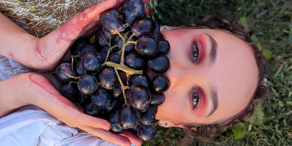 The Bacchae - A teenager's face, beautifully made up, with a background of grass. They have a beard made of rad grapes, which they hold in place with grape-stained hands.