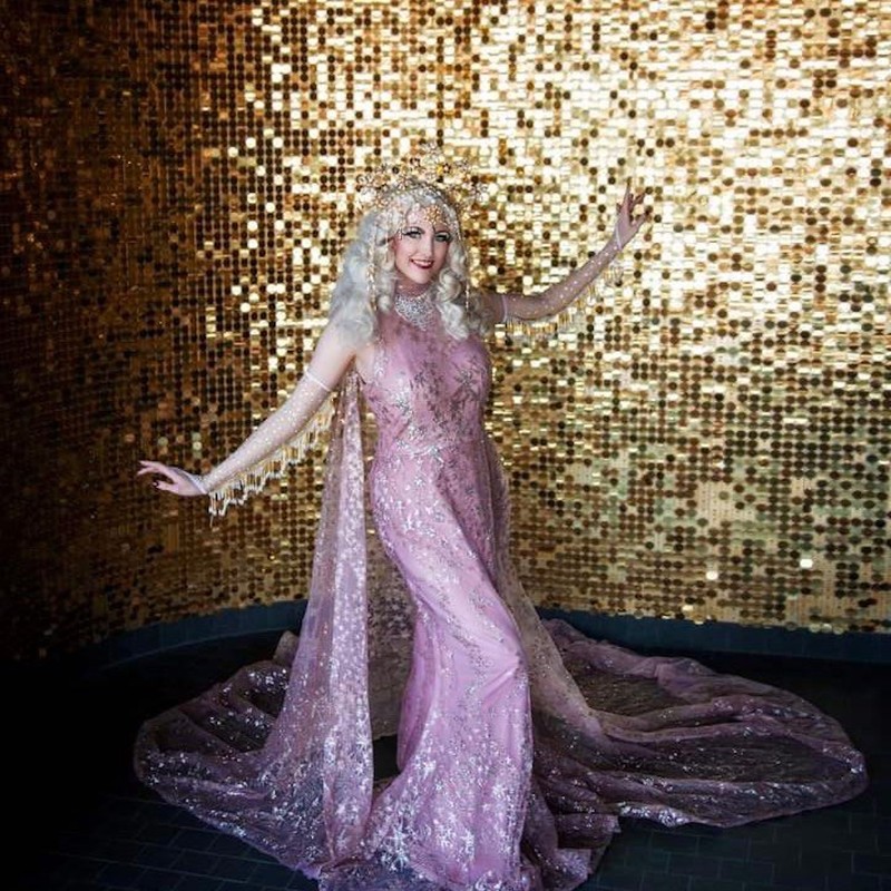 Deco Dolls Burlesque - Blonde burlesque performer standing in front of gold background, wearing long pink dress with glitter stars over the fabric