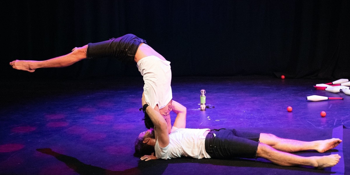 Simon is being held by Ben on his shoulders while he does a flexible back bending handstand. Ben's hands are on Simon's shoulders while he is lying down on the floor.