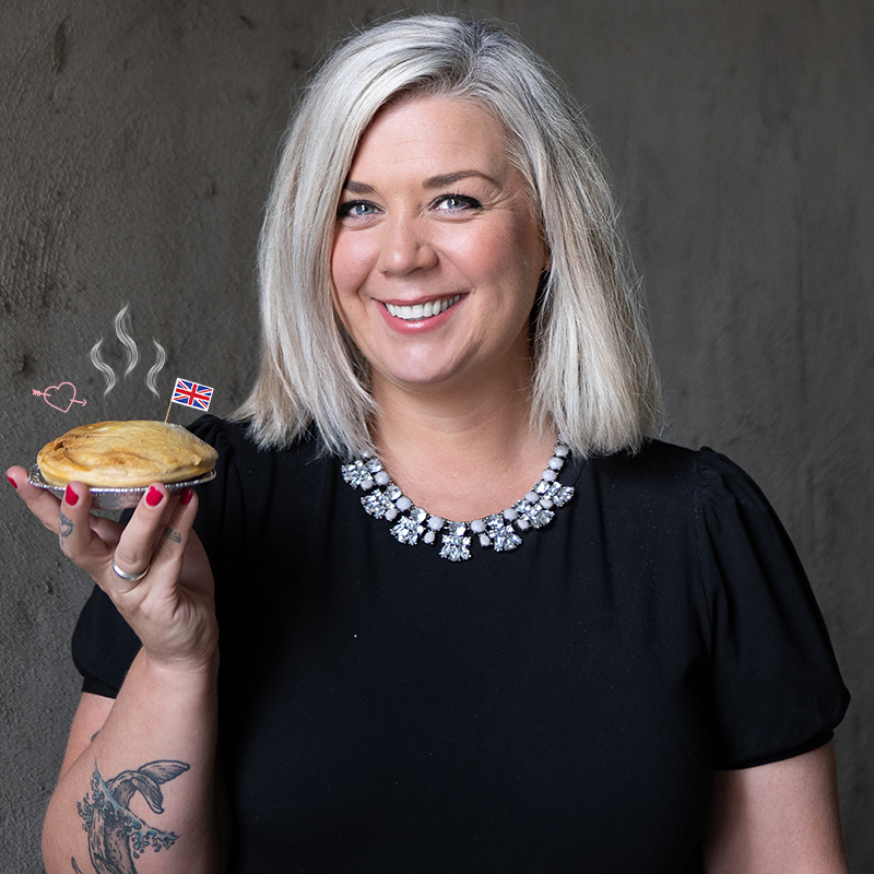 Image of Comedian, Nicky Wilkinson, smiling, wearing a black top and an extravagant diamond necklace. She is holding a hot Steak pie with a British flag stuck in it and steam coming from the top.