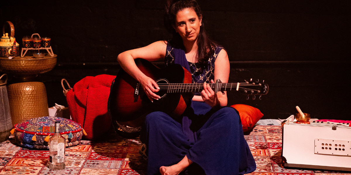 Performer, Almitra, a Pakistani woman living in Australia, sits cross-legged on the floor with a guitar. The floor is covered with lush colourful rugs.