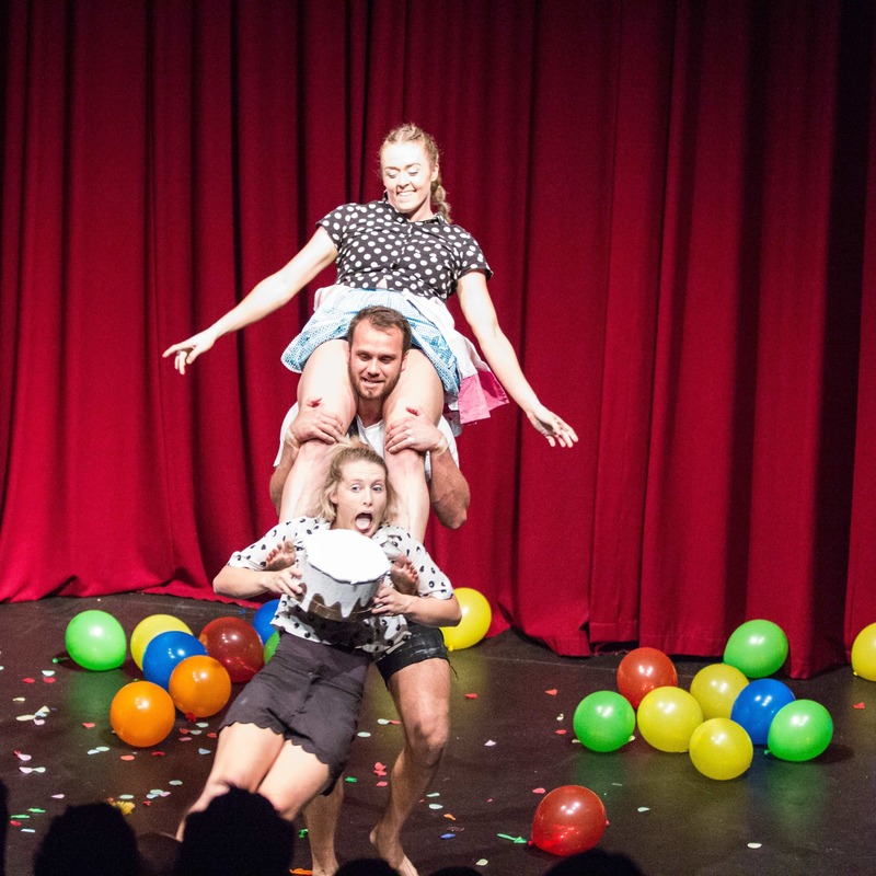 Three people are stacked on top of each other. The person in the forefront is holding a cake and has their tongue out. There is a red curtain in the background and balloons all over the ground.