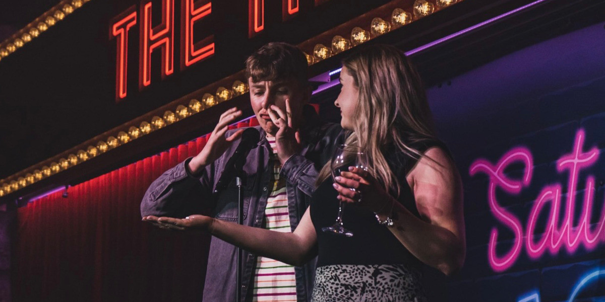 A young man and woman on stage. The young man is snorting something up his nose.