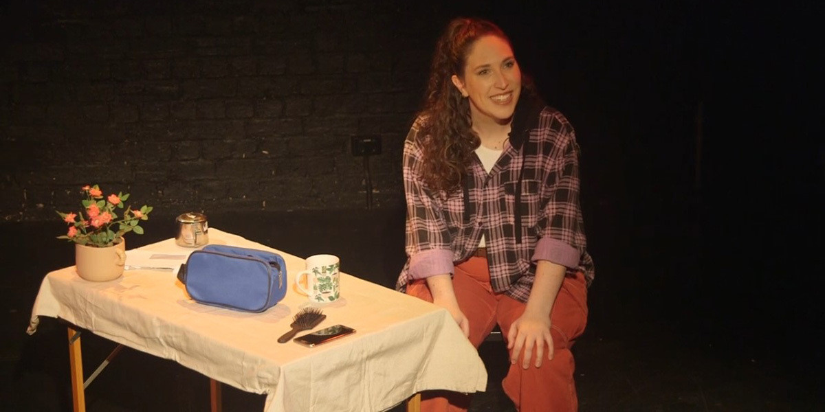 A happy-looking, pale-skinned woman with brown curly hair wearing a purple flannelette shirt and orange pants sits on a chair next to wooden table with a pot plant, hair brush, coffee mug and phone on it.