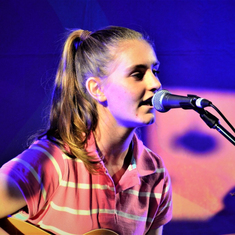 A close up image of Poppy singing. She wears a pink tshirt with thin white, horizontal stripes. She holds a guitar in her lap and a microphone is close to her face. The background is blue in colour.