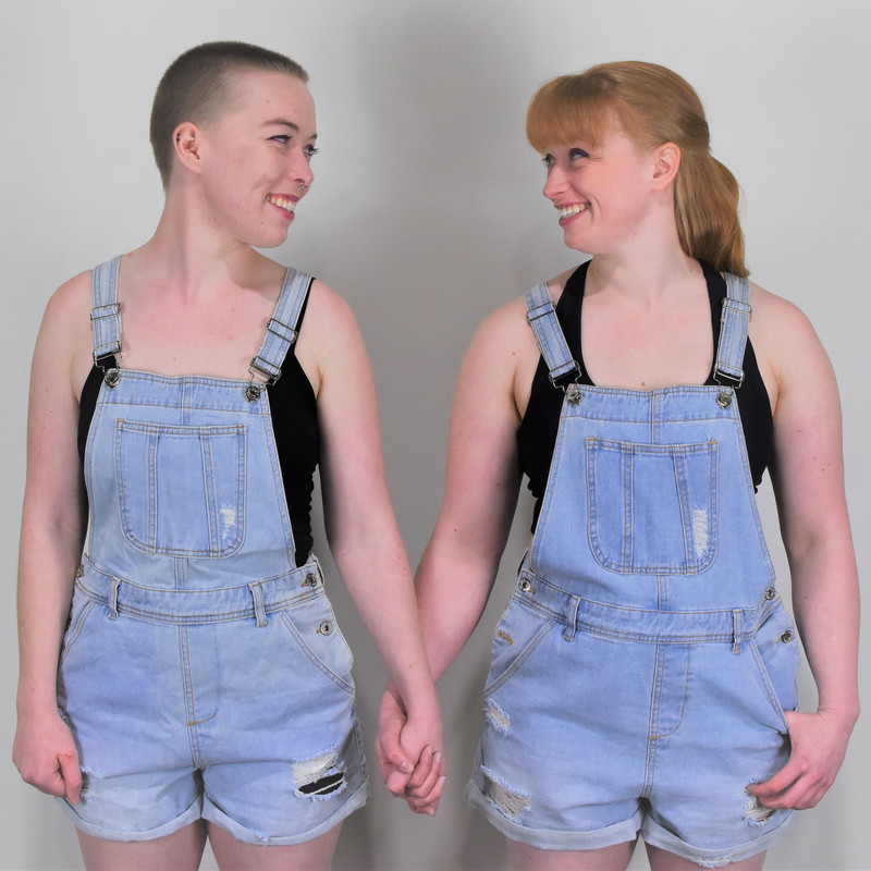 Zoe and Alex wearing light blue denim overalls, holding hands and smiling at each other. The background is white.