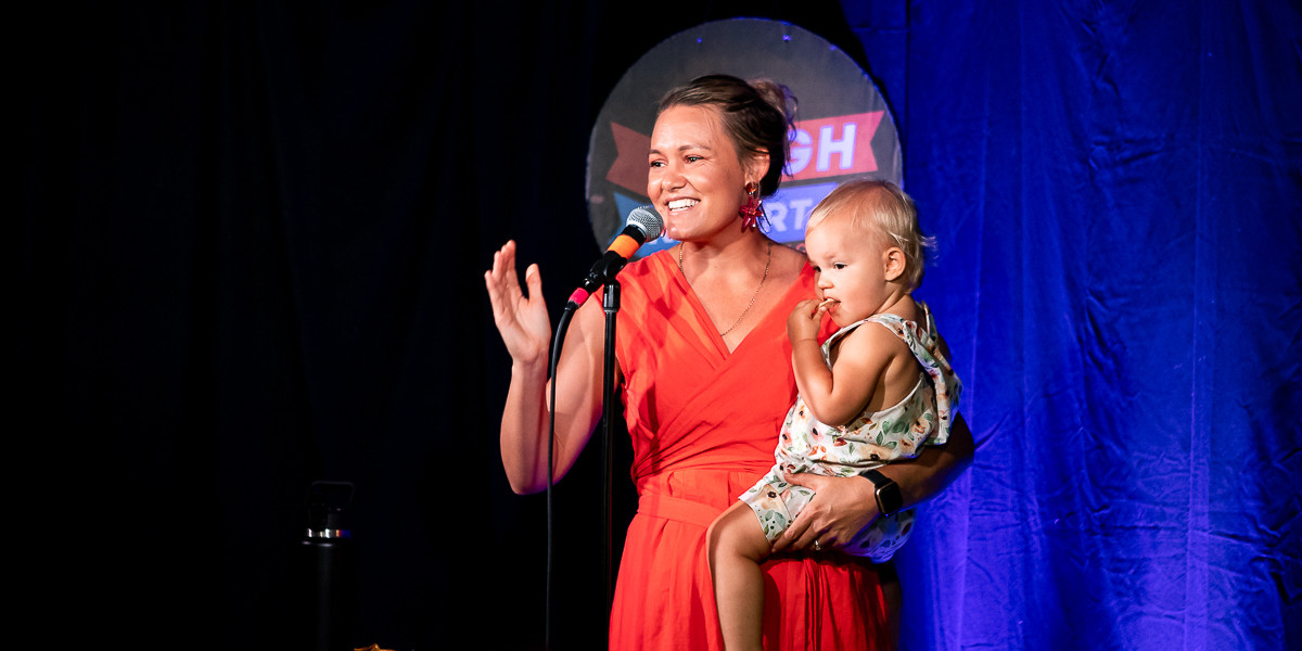 Amy Hetherington: BYO Baby - woman holding baby on stage in front of microphone