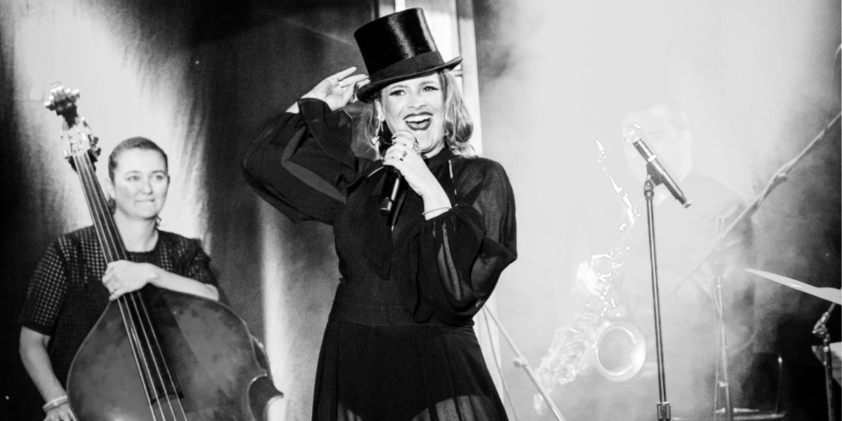 A woman is on stage in a late-night club with a top hat and a huge smile