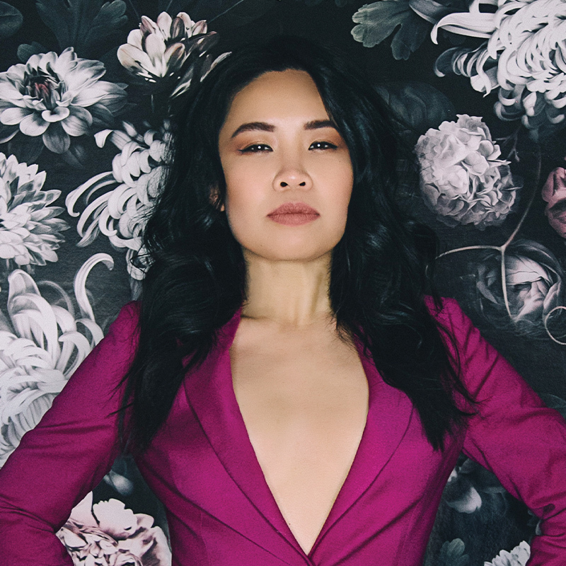 A woman (Diana Nguyen) with dark curly hair, v neck pink top with a serious face. There is a black, white and pink floral background.