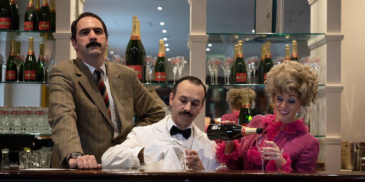 Three performers standing behind a bar. Basil looks angrily at the camera whilst Manuel polishes some glassware and Sybil pours herself some champagne.
