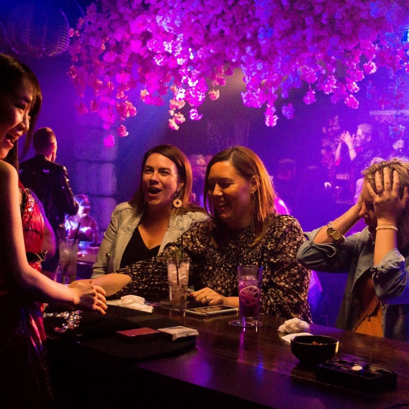 Three women look amazed as a Maho person behind a bar entertains them.