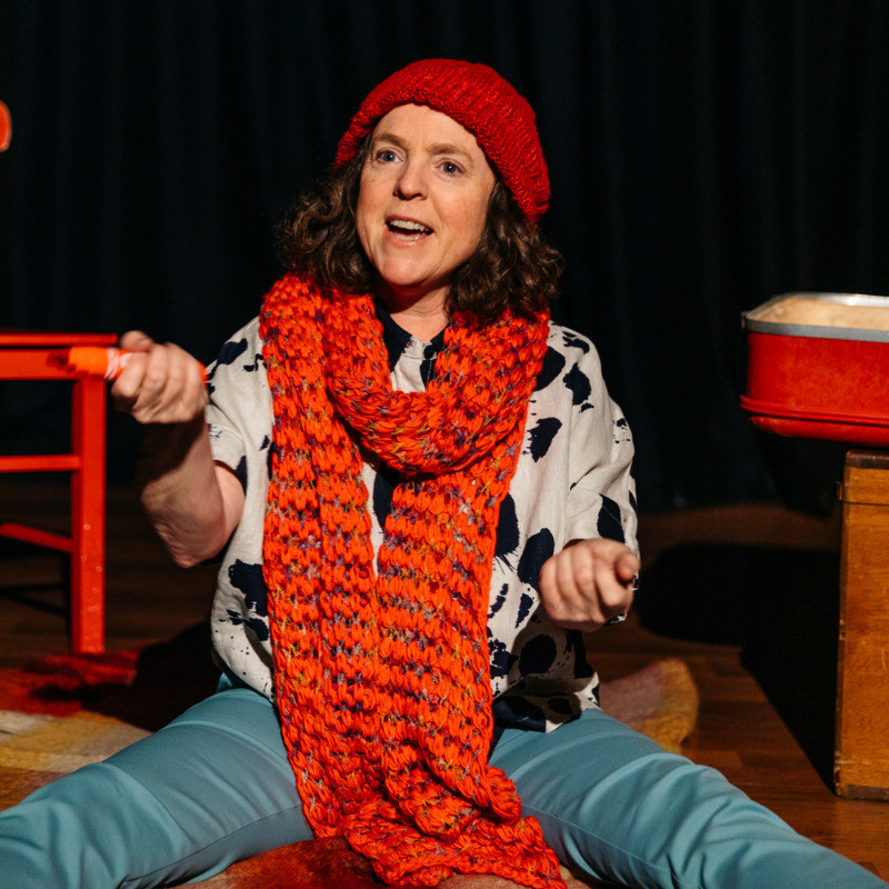 Caroline sits on the floor of a stage performing her show with a red chair and red suitcase alongside her as props. She is wearing a red knitted beanie and a dark orange-red woollen scarf and in her hand is an orange texta.