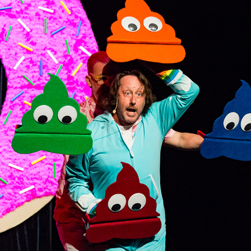 A man in a pale blue onsie is surrounded by 4 poop puppets — 1 orange, 1 green, 1 blue and 1 red. A large pink doughnut stands behind him.