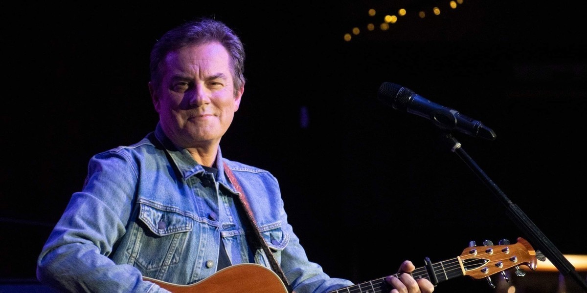 The Long and Winding Road - Close up of a man on a stage in a denim jacket holding an acoustic guitar, while looking directly into the camera.