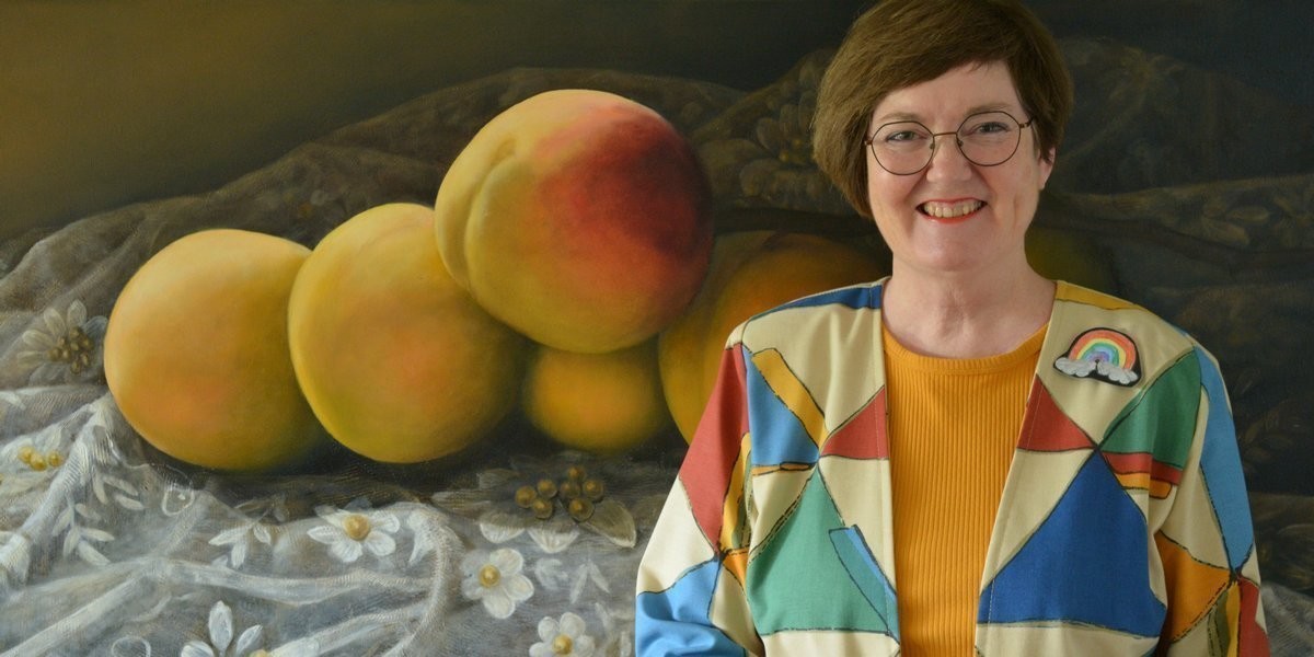 If Fabrics Could Speak... - Artist Catherine Fitz-Gerald, in a colourful geometric patterned coat, stands smiling in front of her large painting of enormous apricots on beaded white lace.