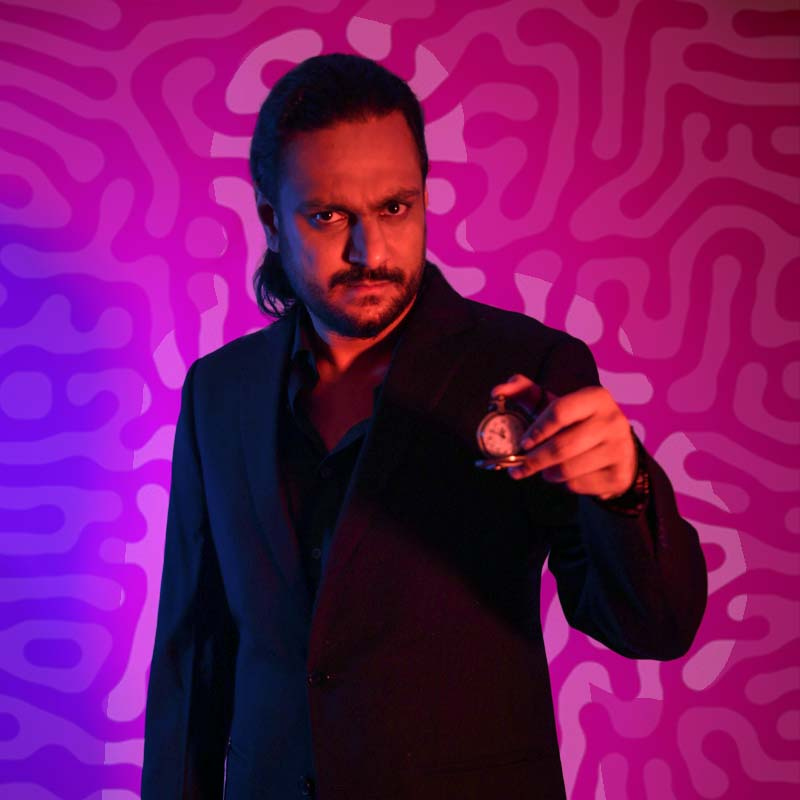 Performer, wearing a black suit, standing in front of a purple textured backdrop, holding a pocket watch, opened and facing the camera