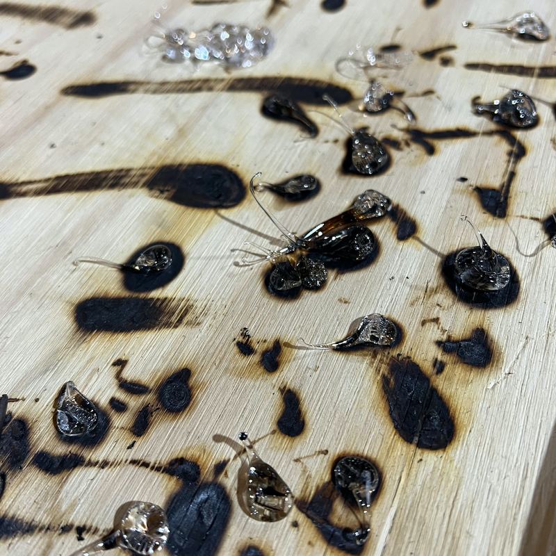 Find That Pace - A light coloured piece of wood with drops of glass that have left burnt marks on the wood.