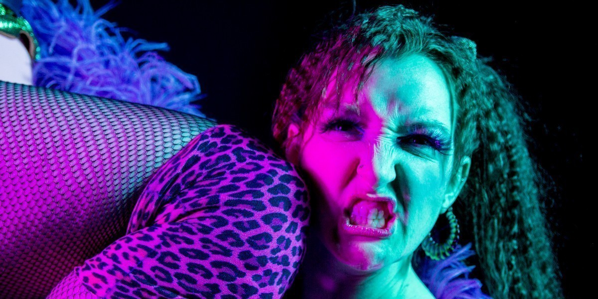 A burlesque wrestler grimaces at the camera while being kneed in the face. The knee is covered with a pink leopard print knee pad and fishnet stockings. They are framed by a purple ostrich feather boa, and are lit by green and pink lighting against a black background.