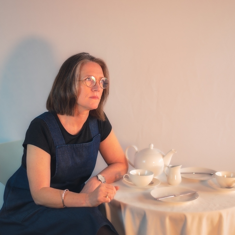 Where To From Here? - Tracy sits at a coffee table looking into the distance. The coffee table is covered in a white tablecloth and is set with a white teaset. The tone of the image is soft white. Tracy is wearing a denim pinafore with a black shirt underneath.