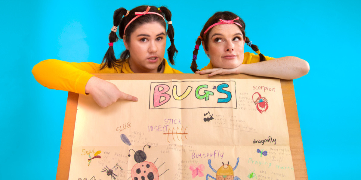 Two white women wearing yellow school polo shirts and with their hair in nostalgic early 2000s hairstyles (including Supre head bands) point proudly at their 'Bugs' school project.
