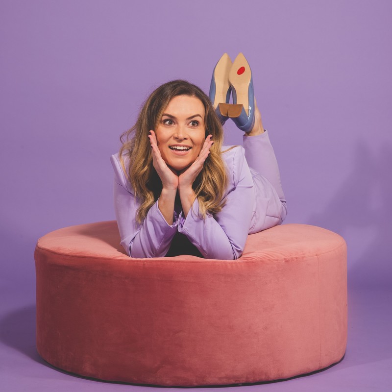 A brown-haired woman in a lavender power suit is lying on her stomach on a pink lounge posing for the camera. Her feet are crossed behind her and a bright red Sale sticker is visible on the sole of her shoe.
