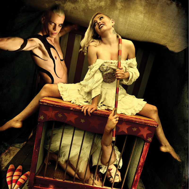 Circus themed, a man stands to the left with arms outreached smirking at the camera. He has a large black tattoo wrapping is arm and body. In the centre a woman sits on top of a wooden cage painted with stars, inside the cage is another woman. The woman in the cage is holding a red and white striped cane out of the bars with the crook around the woman on top's neck.
