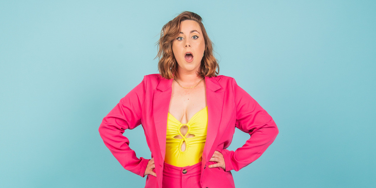 Kate Dolan: A Different Kind of Unhinged - Kate stand front on staring into lense of camera with a shocked expression. She is wearing a hot pink suit with her hands on her hips.
