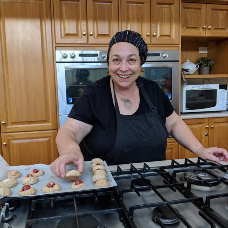 A woman is wearing a black apron and chef hat. She is standing in a home kitchen with a big smile on her face. She has a tray of biscuits in front of her, she is offering the camera one.