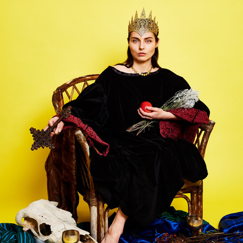 A woman with long dark hair sits in a cane chair surrounded by props. A sheep skull and a gold chalice by her right foot, a red apple in her left hand, an ornate wooden cross in her right hand, some flowers on her lap, and gold crown on her head. The background is bright yellow, she wears a black tunic.