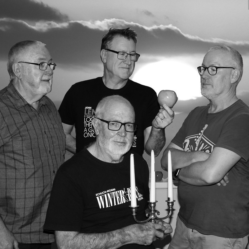Duelling Duos - A black and white photo of four older men, three standing and one sitting in the centre front, he holds a three pronged candelabra. The one in at the back in the centre holds an apple. They all have short hair and wear glasses. The background is a cloudy sky.