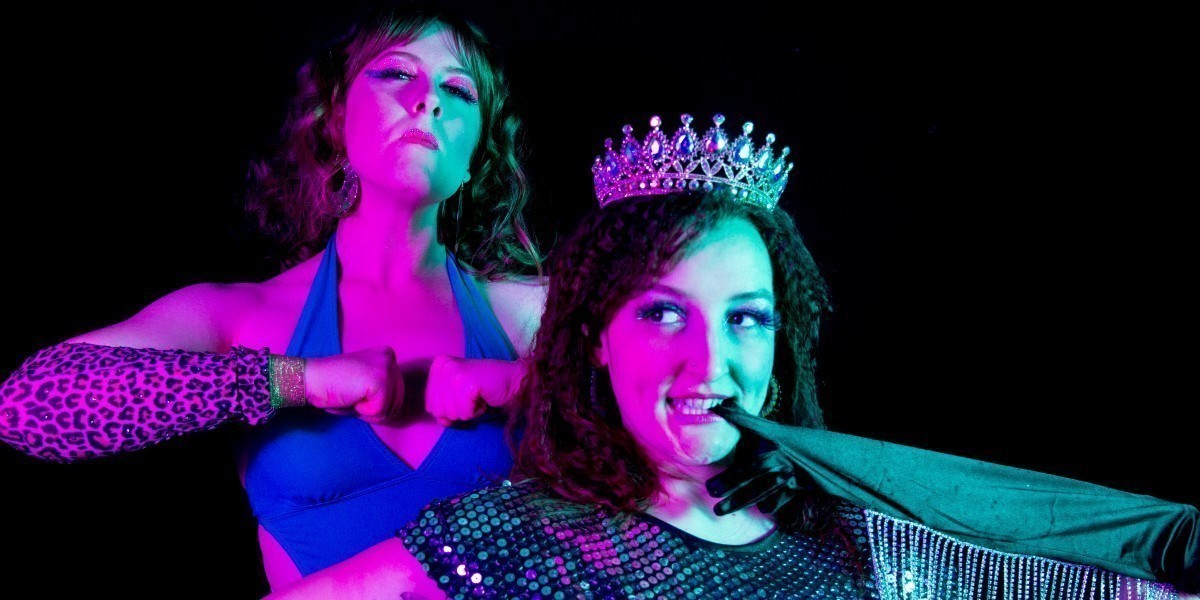 A burlesque performer in a silver sequined gown and crown poses cheekily in the foreground. She’s biting a black satin glove between her teeth and has her other hand on her hip. Another stands behind her wearing an 80’s wrestler style green leotard and pink and gold gauntlets. She has her head tilted back and fists clenched in front of her in a power pose. They are lit by green and pink lighting against a black background.