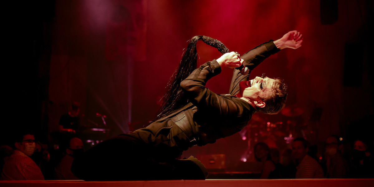Side profile of man wearing all black on a stage. He is on his knees, leaning backwards towards the ground as he sings into a black microphone with long black tassels at the end. He has on drag makeup with false eyelashes. Surrounding him is ambient red lighting and haze, with out of focus audience members in the background.
