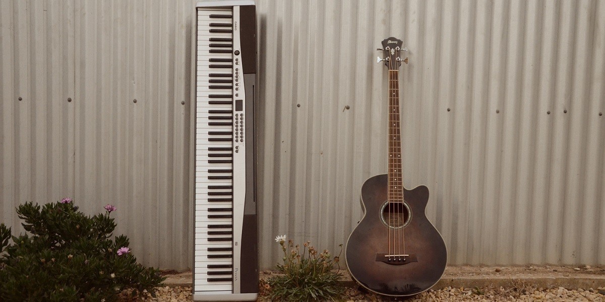 A keyboard and an acoustic bass guitar lean against a corrugated iron shed.