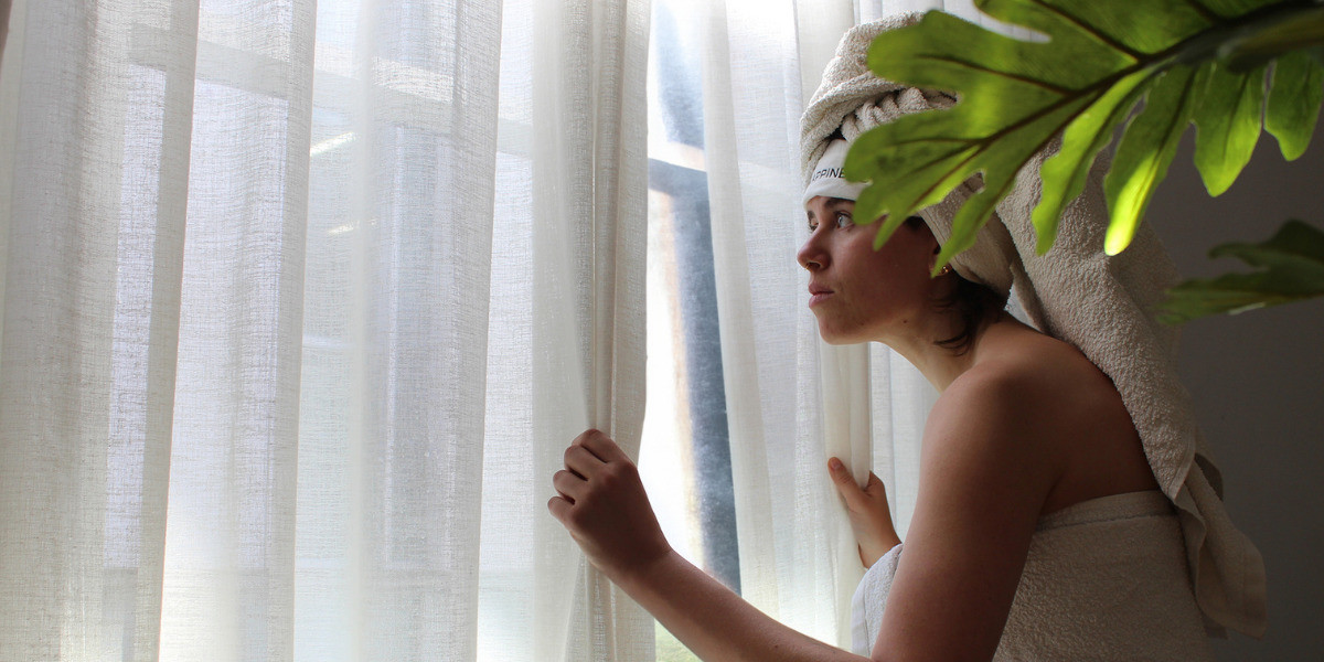 MASSAGE THERAPY - A woman wearing a headband with 'HAPPINESS' printed on it, a bath towel, and a head towel, stands between flowing white curtains. She raises her hands, moving the curtains, while looking up and to the left. Sunlight from a right-side window illuminates her face, with steam in the background.