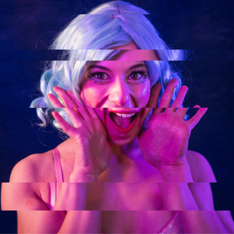 A performer in a light blue wig and doll-like makeup smiles too widely at the camera. Her hands are beside her face as if to say 'tah-dah!' The image is distorted like it's been cut into 5 pieces.