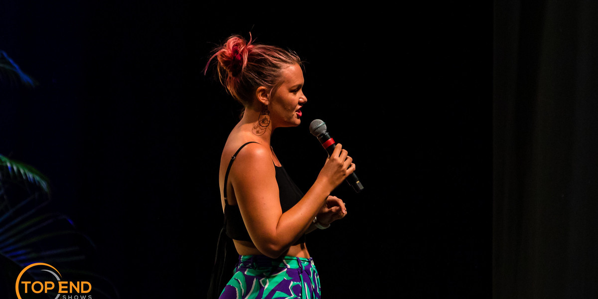 Woman stands on stage with a microphone. Black background. She is performing stand up comedy
