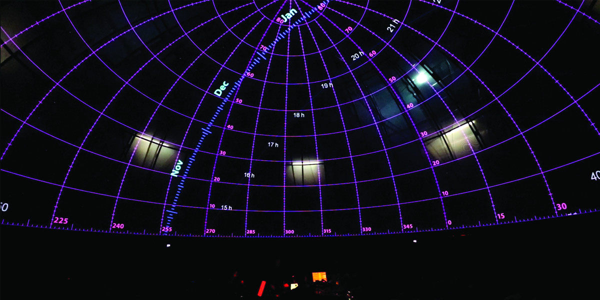 Musicians sit beneath a domed planetarium ceiling, silhouetted by the glow of the iPads they are reading from. The ceiling features graphic datelines and degrees in purple, green, blue and pink against a dark backdrop.