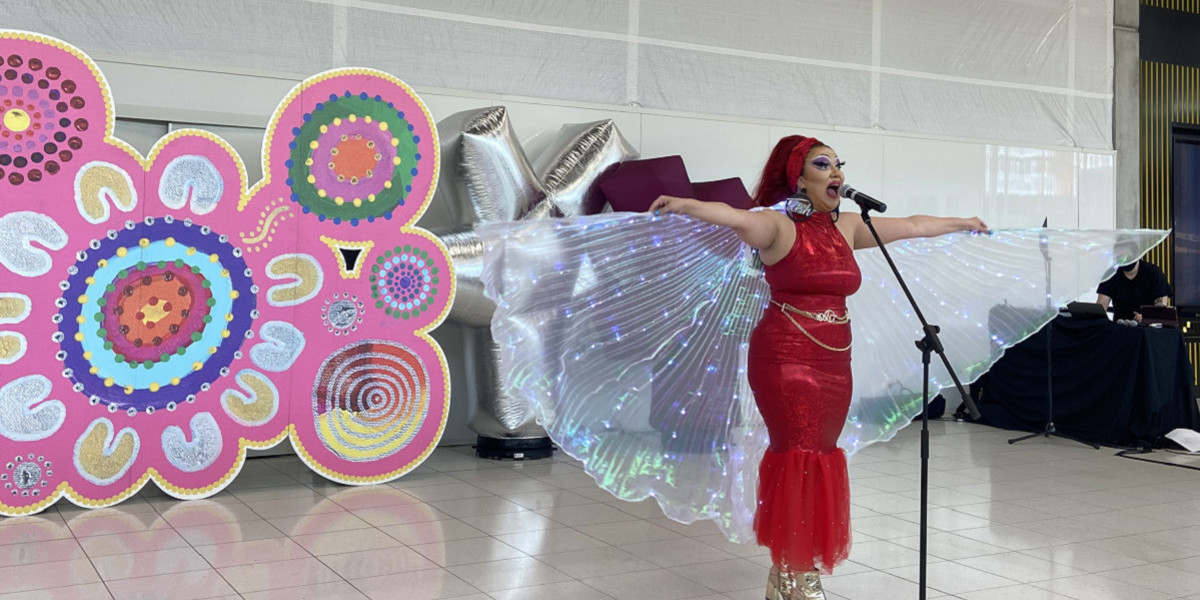 An artist wearing a sparkly red dress stands in front of a microphone with light-up wings spread. She stands in front of a bright pink backdrop in the airport.