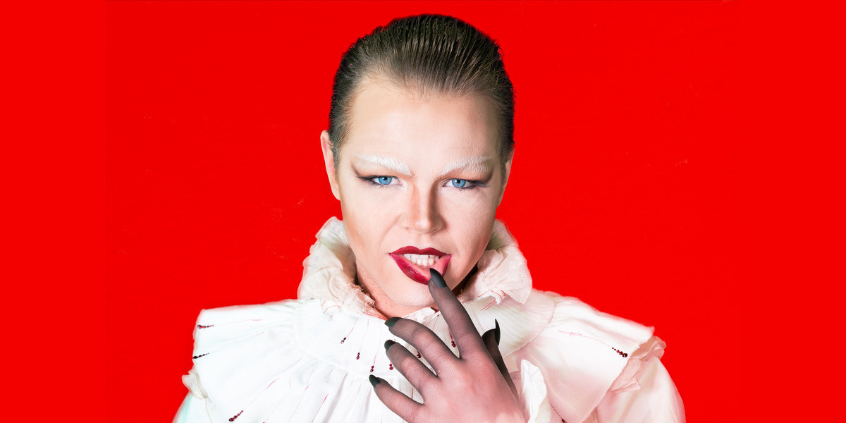 Florian Wild, a caucasian man with blue eyes and brown hair, looks directly at the camera. He has drag queen makeup on, with white eyebrows and a red lip. He is dressed in a white blouse and a tall white vampire collar with red diamonte accents like blood droplets. He has black nails on his hands.
