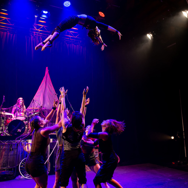 A group of acrobats throw a woman high into the air while she flips upside down, their arms are out ready to catch her. They are wearing torn clothing and behind them is piles of broken furniture and tools. In the background a female musician plays the drums and sings on an elevated platform.