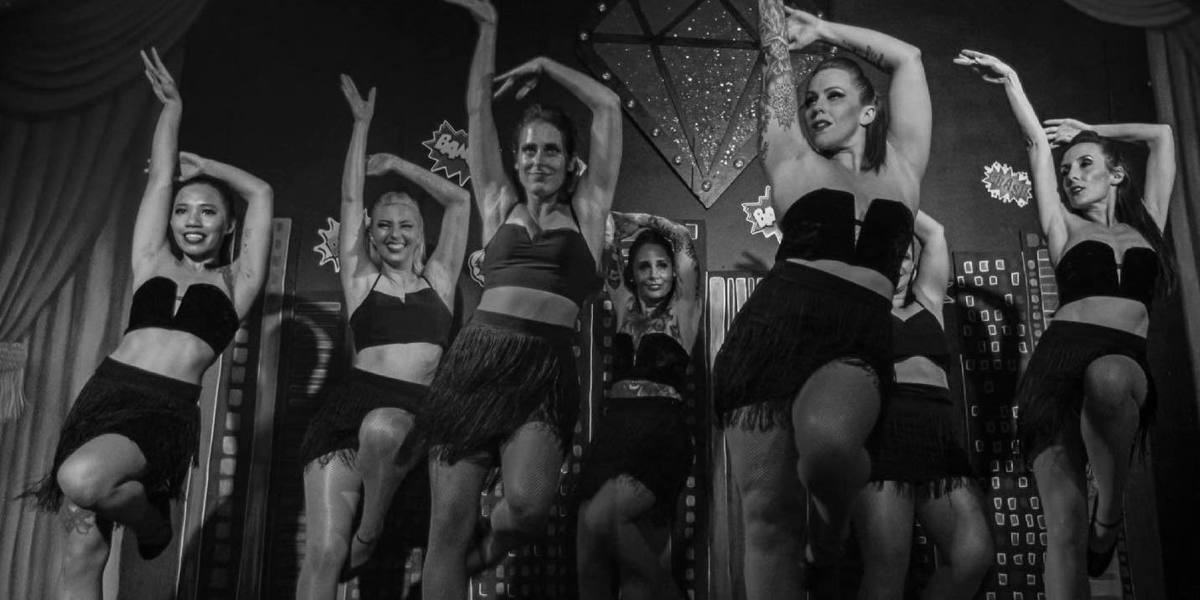 a black and white image depicts a group of dancers standing in a post together in a Jazz style balanced on one leg