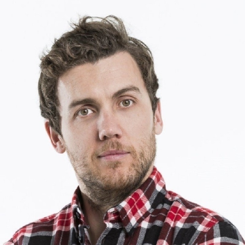 A headshot photo of a man with short brown hair and a brown beard staring at the camera. He is wearing a red, black and white checked shirt. The background is white.