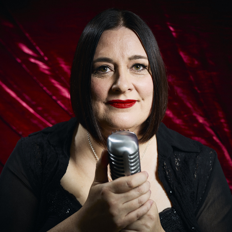Alexandra Frost -Cabaret Singer/Writer/Producer of cabaret show "Hear My Voice" for Adelaide Fringe, 2023, dressed in black with red velvet background holding vintage mic