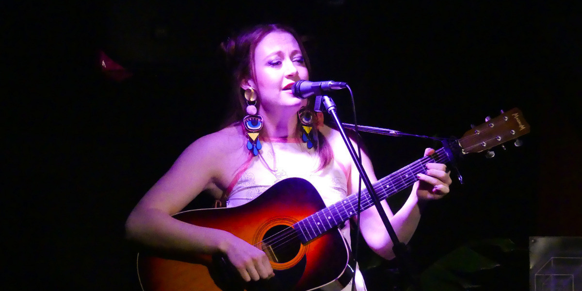 A mid shot of Steph playing a guitar and singing into a microphone. She is wearing a pink strappy tank top and brightly coloured earings.