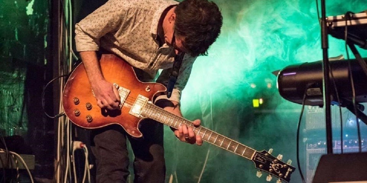 A photo of a person performing on stage. They are leaning over their guitar. The background is illuminated with green lights and smoke.