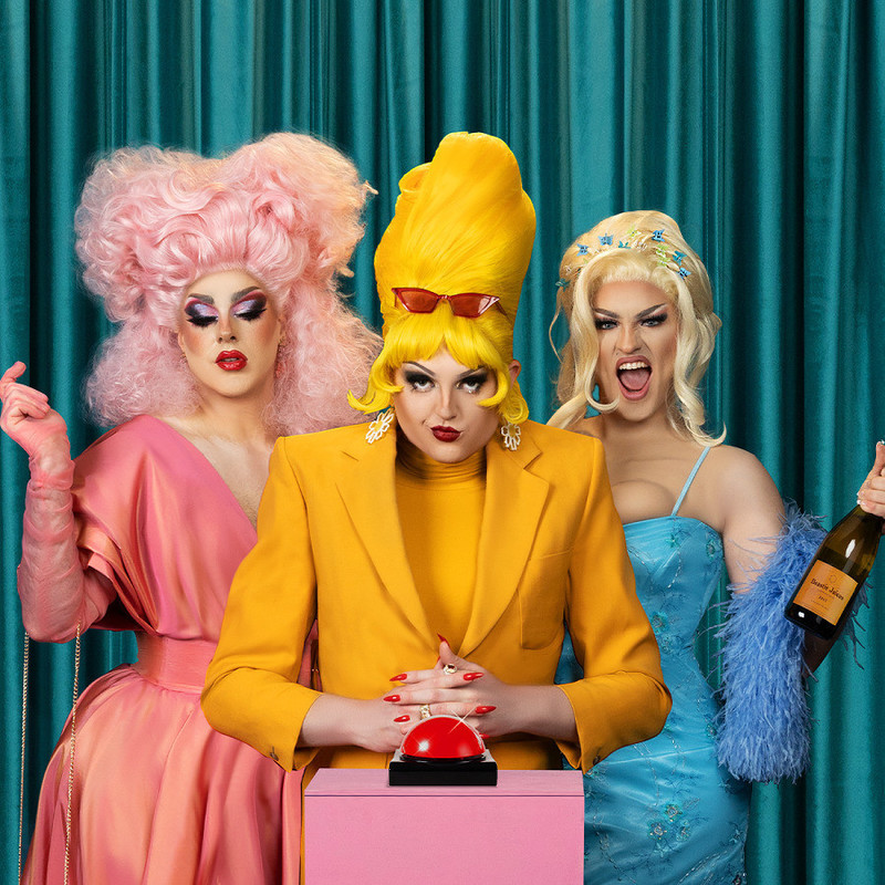 The Beastie Girls are three drag performers standing together in front of a pink background. Lazy Suzan is wearing a yellow dress and a tall yellow wig, Zelda Moon is wearing a pink dress and a sleek pink wig and Benign Girl is wearing a blue dress and a sexy blonde wig.