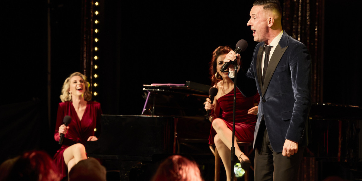 Two women sit by the piano laughing, while a man stands talking into microphone to audience