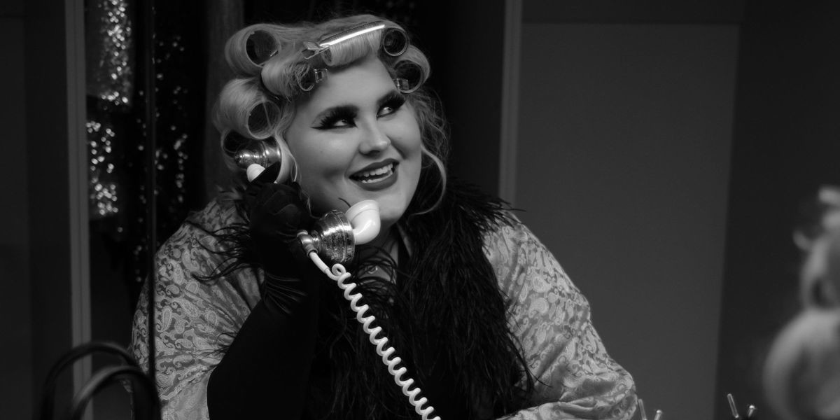 Charlee Watt in a vintage style black and white shot, sits in front of a mirror with her hair in vintage curlers and clips. There are vintage handbags, jewlery and accessories on the dresser. Charlee is speaking into a vintage telephone about to burst into laughter.