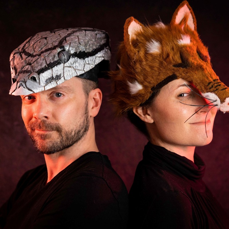 Two performers standing back to back with heads slightly turned facing the camera. One performer wearing a fox costume for her head and other performer wearing a snake one.