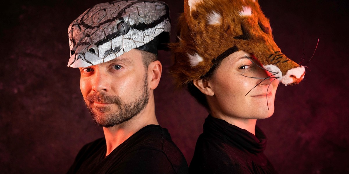 Two performers standing back to back with heads slightly turned facing the camera. One performer wearing a fox costume for her head and other performer wearing a snake one.